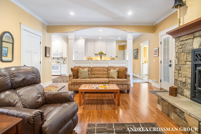 living area featuring a stone fireplace, ornamental molding, light wood-style flooring, and decorative columns