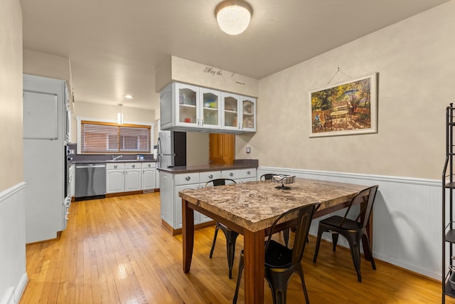 kitchen featuring a wainscoted wall, light wood finished floors, dark countertops, appliances with stainless steel finishes, and glass insert cabinets