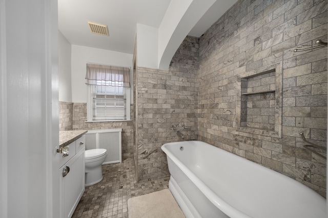 full bath with visible vents, toilet, a soaking tub, vanity, and tile walls