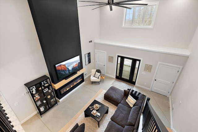 living area featuring a glass covered fireplace, ceiling fan, a towering ceiling, and baseboards