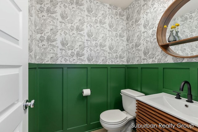 half bathroom with wallpapered walls, wainscoting, toilet, vanity, and a decorative wall