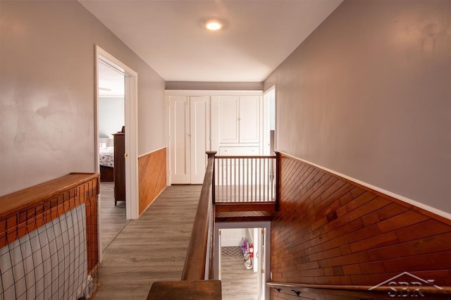 corridor with a wainscoted wall, wood walls, light wood finished floors, and an upstairs landing