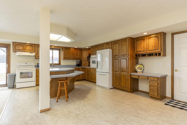 kitchen with under cabinet range hood, white appliances, light floors, brown cabinetry, and a kitchen bar