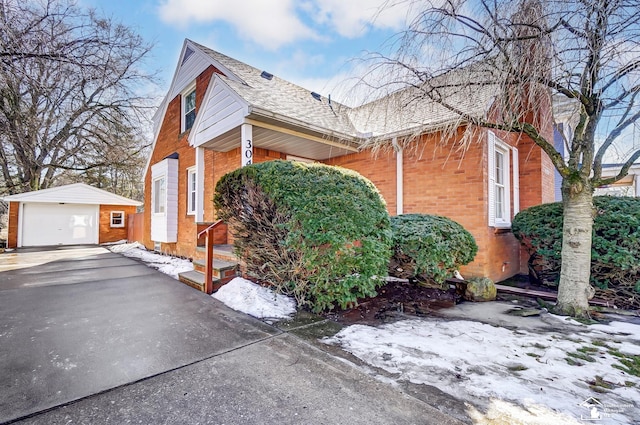 view of snow covered exterior featuring aphalt driveway, a shingled roof, brick siding, an outdoor structure, and a detached garage