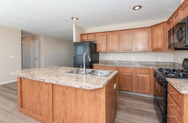 kitchen with a center island with sink, brown cabinetry, light wood-type flooring, black appliances, and a sink