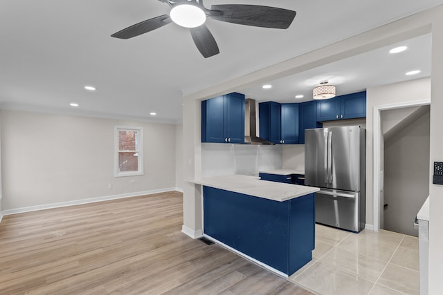 kitchen featuring wall chimney range hood, freestanding refrigerator, a peninsula, light countertops, and blue cabinetry