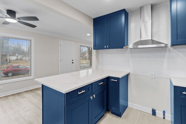 kitchen with blue cabinetry, a peninsula, wall chimney exhaust hood, and light stone countertops