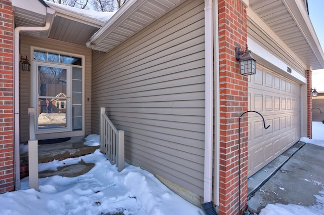 snow covered property entrance with an attached garage and brick siding