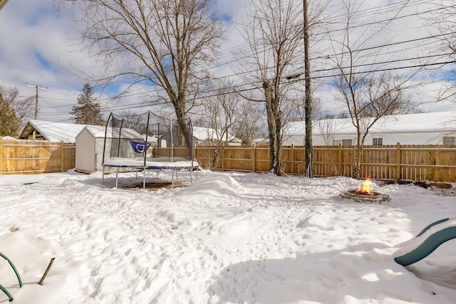 yard covered in snow with a storage shed, an outdoor fire pit, a fenced backyard, an outbuilding, and a trampoline