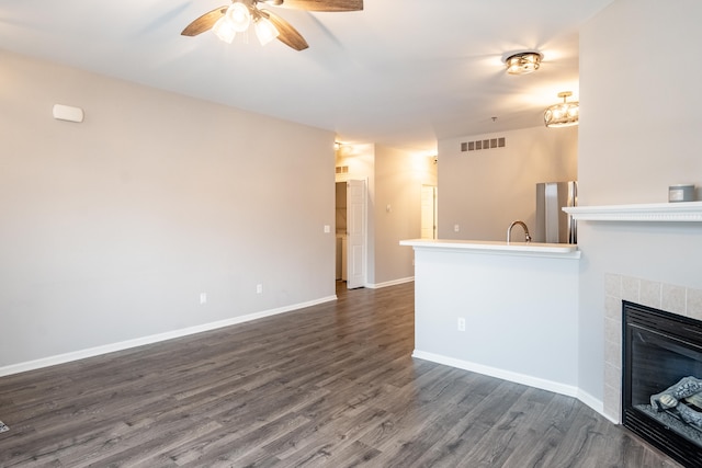 unfurnished living room featuring a tile fireplace, a ceiling fan, visible vents, baseboards, and dark wood-style floors