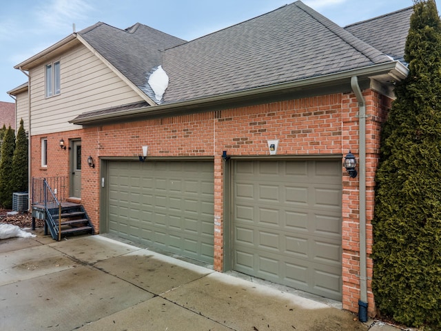 view of side of property with a garage, brick siding, central AC unit, and a shingled roof