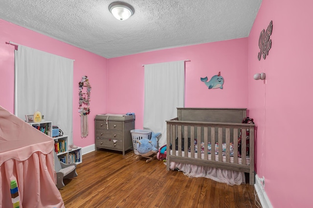 bedroom with a nursery area, a textured ceiling, baseboards, and wood finished floors