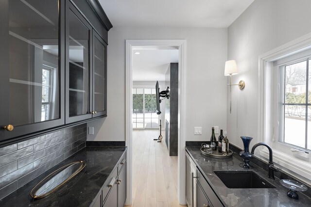 kitchen with light wood-style flooring, a sink, decorative backsplash, dark stone counters, and glass insert cabinets