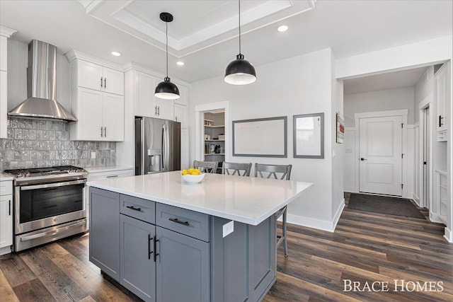 kitchen with a tray ceiling, wall chimney exhaust hood, stainless steel appliances, white cabinetry, and a kitchen breakfast bar