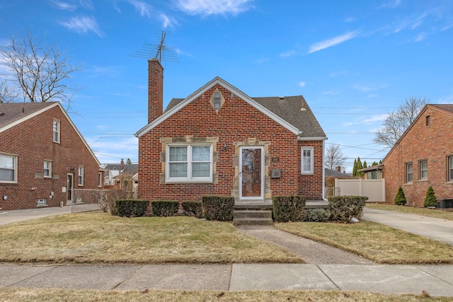 view of front facade featuring brick siding, a chimney, a shingled roof, fence, and a front lawn