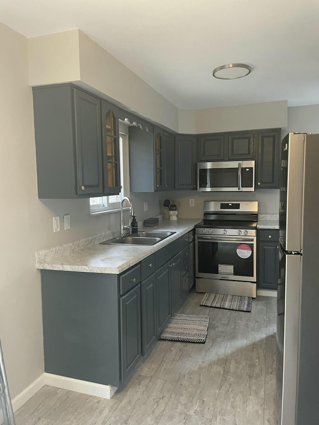 kitchen with light countertops, gray cabinetry, appliances with stainless steel finishes, glass insert cabinets, and a sink