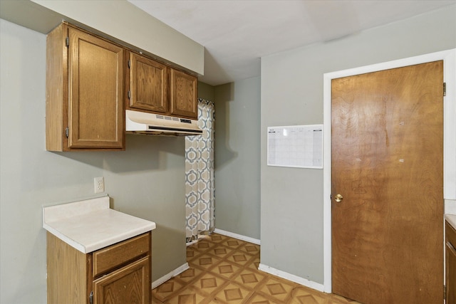 kitchen featuring brown cabinetry, light countertops, under cabinet range hood, and baseboards