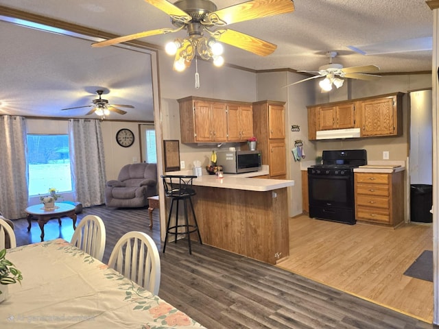 kitchen with black gas range oven, light wood-style flooring, stainless steel microwave, a peninsula, and under cabinet range hood