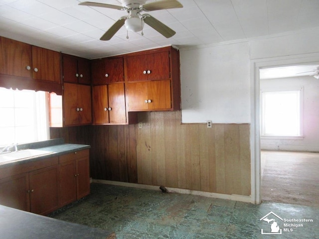 kitchen with a wainscoted wall, brown cabinets, a ceiling fan, a sink, and wood walls