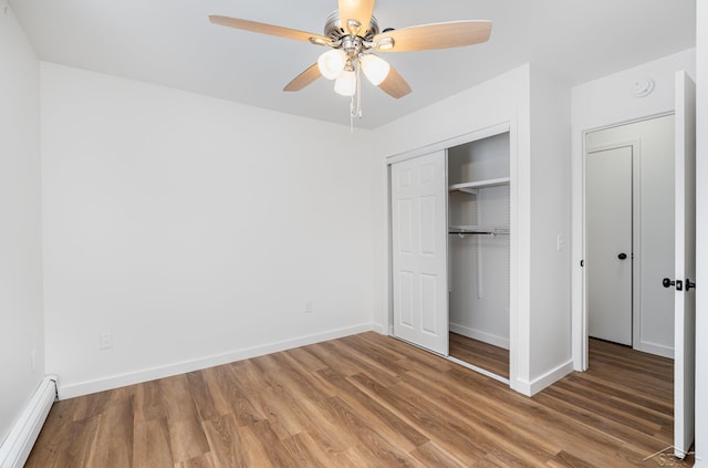 unfurnished bedroom with a closet, a baseboard heating unit, ceiling fan, wood finished floors, and baseboards