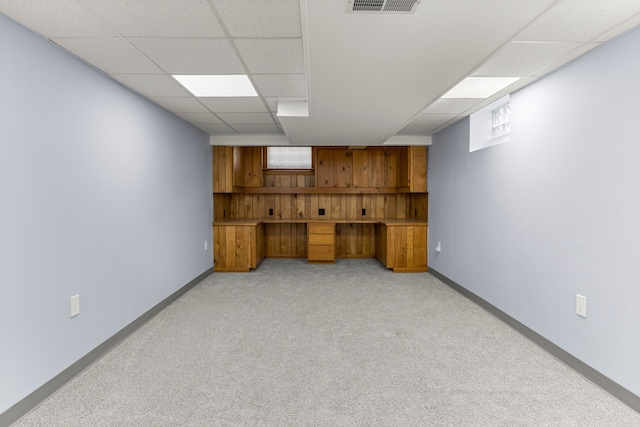 below grade area with visible vents, light carpet, wood walls, a drop ceiling, and baseboards