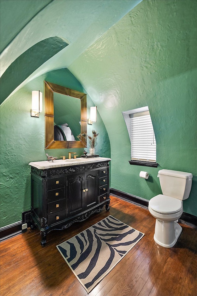 bathroom featuring double vanity, a textured wall, toilet, wood-type flooring, and vaulted ceiling