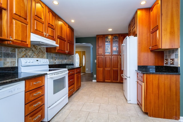 kitchen with white appliances, dark stone countertops, backsplash, and under cabinet range hood