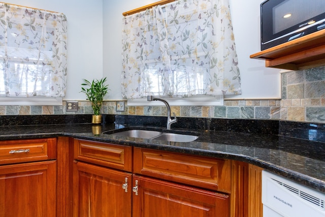 kitchen with brown cabinetry, white dishwasher, a sink, dark stone countertops, and black microwave