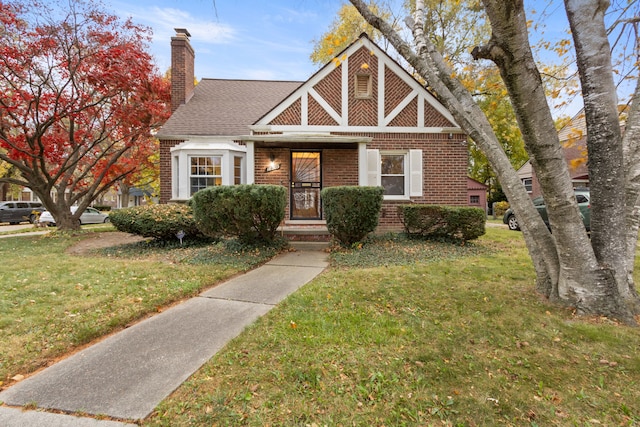 english style home with a shingled roof, a front yard, brick siding, and a chimney