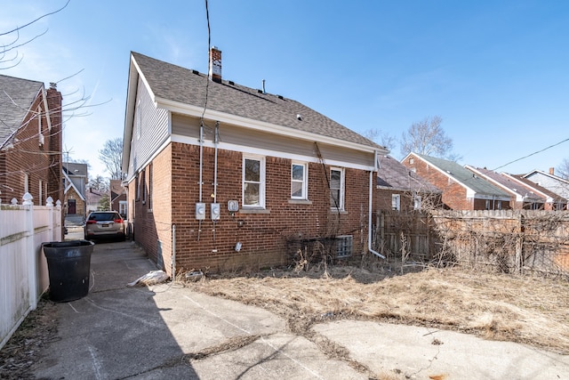 rear view of property featuring brick siding, a chimney, roof with shingles, and fence