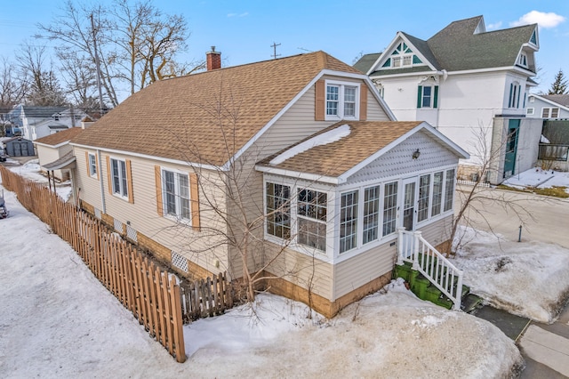 snow covered property featuring a sunroom, a shingled roof, a chimney, and fence