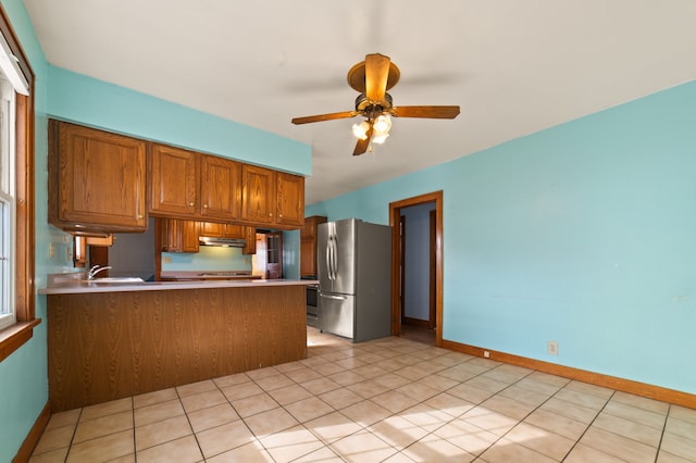 kitchen featuring under cabinet range hood, a peninsula, a ceiling fan, freestanding refrigerator, and brown cabinetry