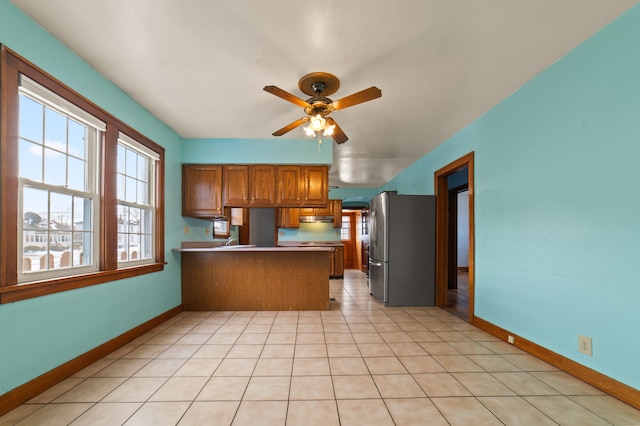 kitchen with brown cabinetry, a ceiling fan, freestanding refrigerator, a peninsula, and light tile patterned flooring