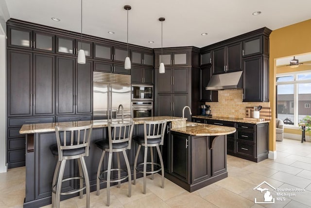 kitchen featuring built in appliances, under cabinet range hood, a breakfast bar, backsplash, and light stone countertops