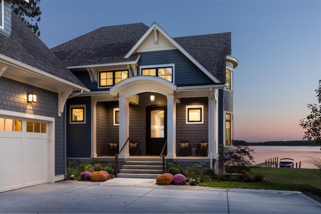exterior entry at dusk featuring a garage, driveway, a shingled roof, a water view, and a porch