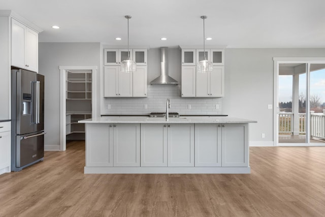 kitchen featuring stainless steel fridge with ice dispenser, backsplash, an island with sink, light wood-type flooring, and wall chimney exhaust hood