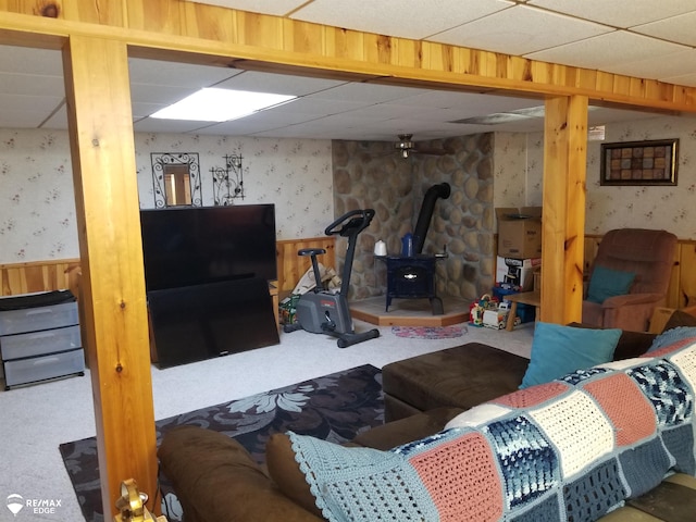 living room featuring a wainscoted wall, a drop ceiling, carpet, wallpapered walls, and a wood stove