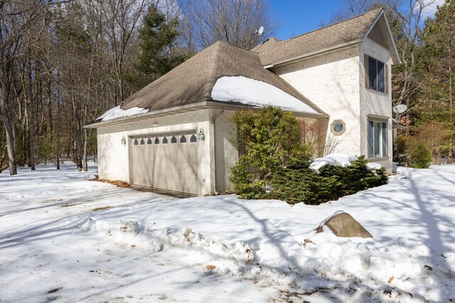 view of snow covered exterior featuring a shingled roof, an attached garage, and stucco siding