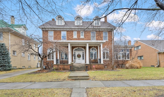 view of front of home with a porch, brick siding, a shingled roof, a front lawn, and a chimney