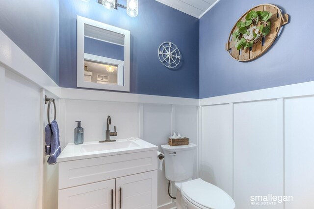 half bathroom featuring a wainscoted wall, vanity, toilet, and a decorative wall