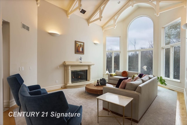 living area featuring beam ceiling, visible vents, a glass covered fireplace, high vaulted ceiling, and baseboards