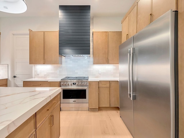 kitchen featuring premium appliances, light brown cabinets, light wood-type flooring, decorative backsplash, and wall chimney exhaust hood