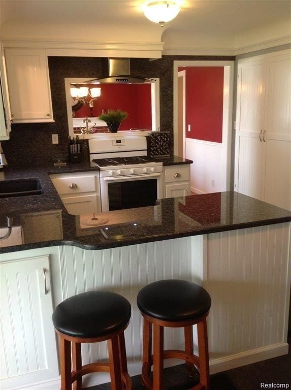 kitchen with white cabinetry, a sink, wall chimney range hood, and white gas range oven