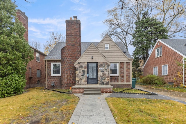 english style home featuring stone siding, a chimney, roof with shingles, a front lawn, and brick siding