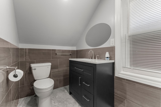 bathroom featuring a wainscoted wall, tile walls, lofted ceiling, toilet, and vanity