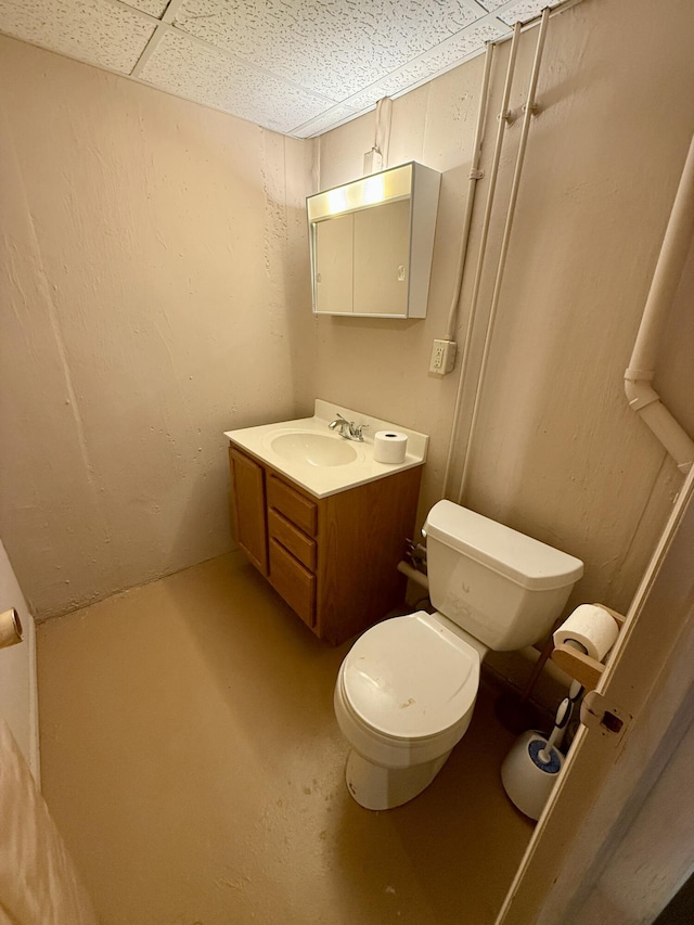 half bathroom featuring concrete flooring, a textured wall, a drop ceiling, toilet, and vanity