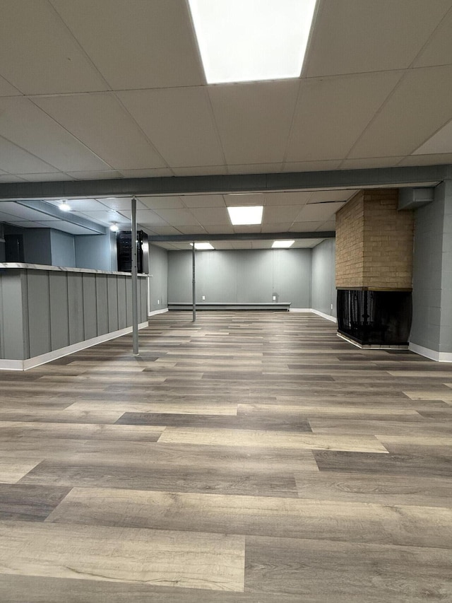 finished basement featuring a brick fireplace, a drop ceiling, baseboards, and wood finished floors