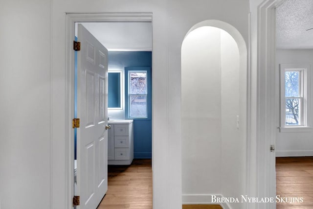 hall featuring light wood-type flooring, baseboards, arched walkways, and a textured ceiling