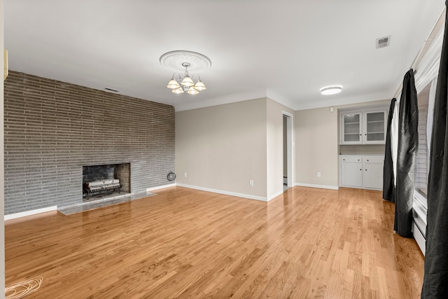 unfurnished living room with a brick fireplace, baseboards, visible vents, light wood-style floors, and a chandelier