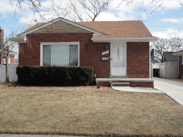 bungalow-style house with a shingled roof, a front yard, brick siding, and fence